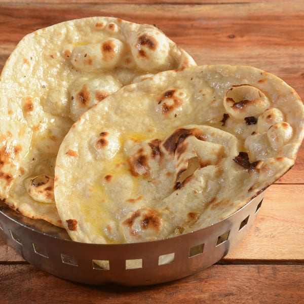 Golden-brown Indian naan bread, glistening with butter, served in a metal basket on a wooden table.