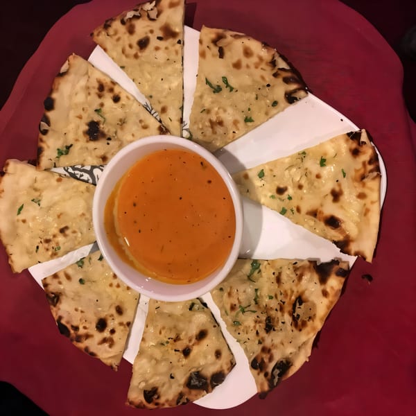 New Taj Mahal's garlic naan bread slices arranged around a bowl of orange dipping sauce on a red tablecloth.