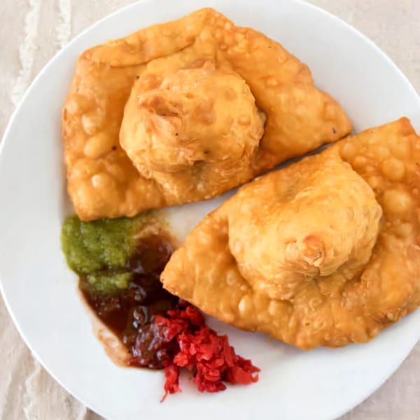 Two golden-brown Indian samosas with green chutney, tamarind sauce, and red pickled onions on a white plate.